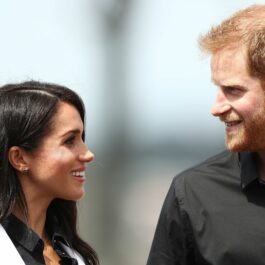 Prințul Harry, îmbrăcat într-o cămașă neagră, îi zâmbește soției sale, ce poartă o cămașă neagră și un sacou alb